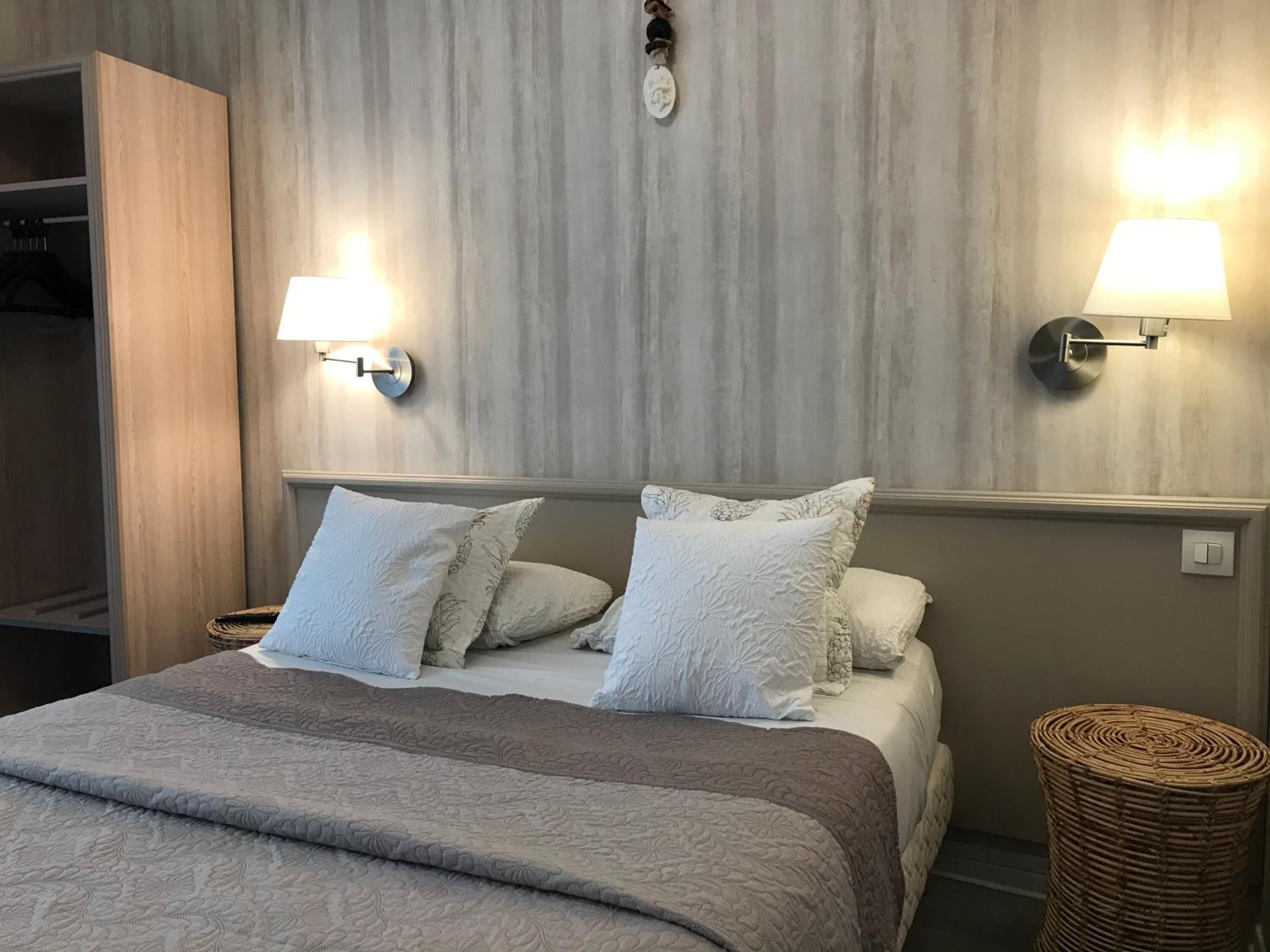 A neatly made bed with a grey quilt and white pillows, two wall-mounted lamps, and a wicker basket beside the bed.