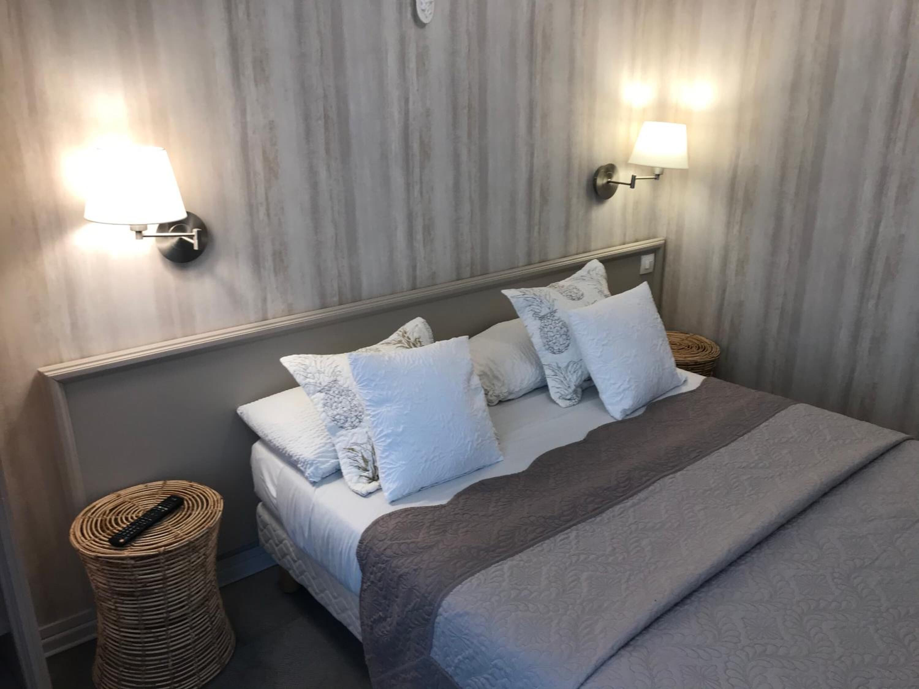 A neatly made bed with a grey blanket and multiple pillows, two wall-mounted lamps, and a woven basket on the floor.