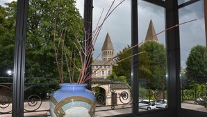 Blick durch ein Fenster mit einer dekorativen Vase auf einem Fensterbrett, einem Balkongeländer und einem Gebäude mit zwei Türmen im Hintergrund.