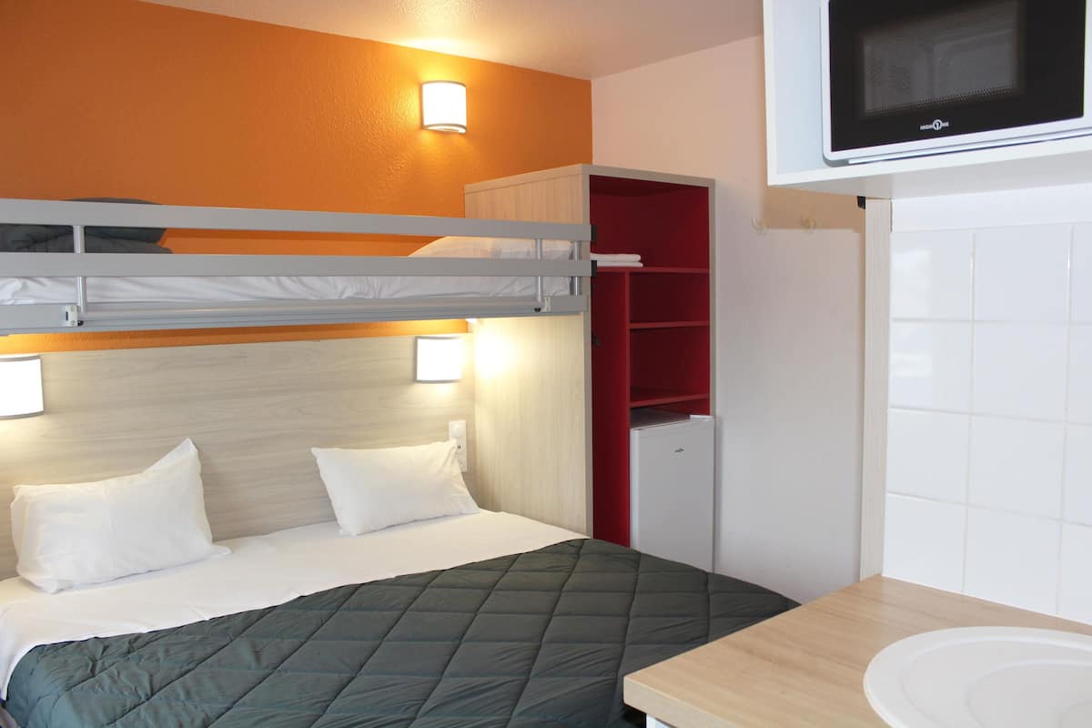 A bunk bed with a green quilt, a white bedside table, a microwave above the bed, and a red storage unit.