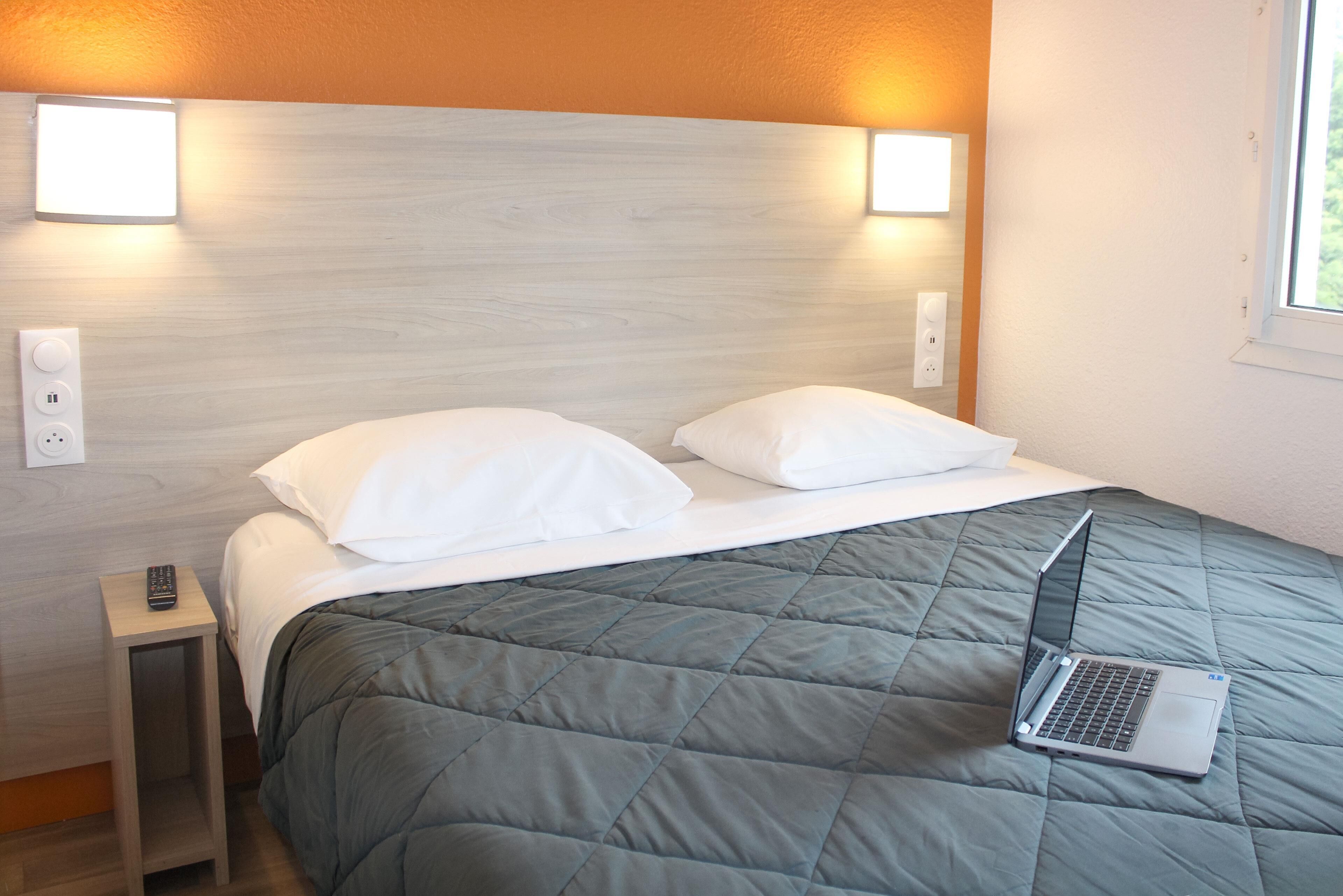 Een net opgemaakt bed met een laptop erop, een nachtkastje met een afstandsbediening en wandlampen.