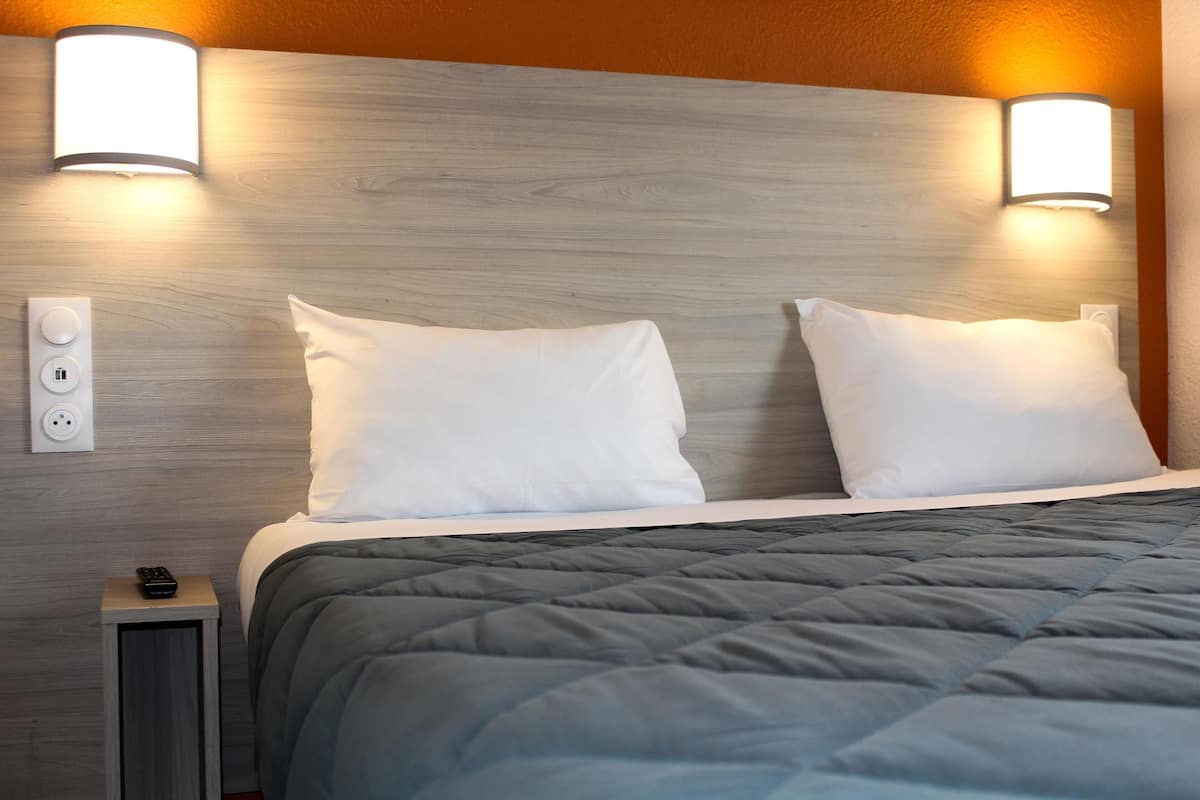 A hotel room with a wooden headboard, two white pillows, a grey quilted bedspread, and a bedside table with a remote control.
