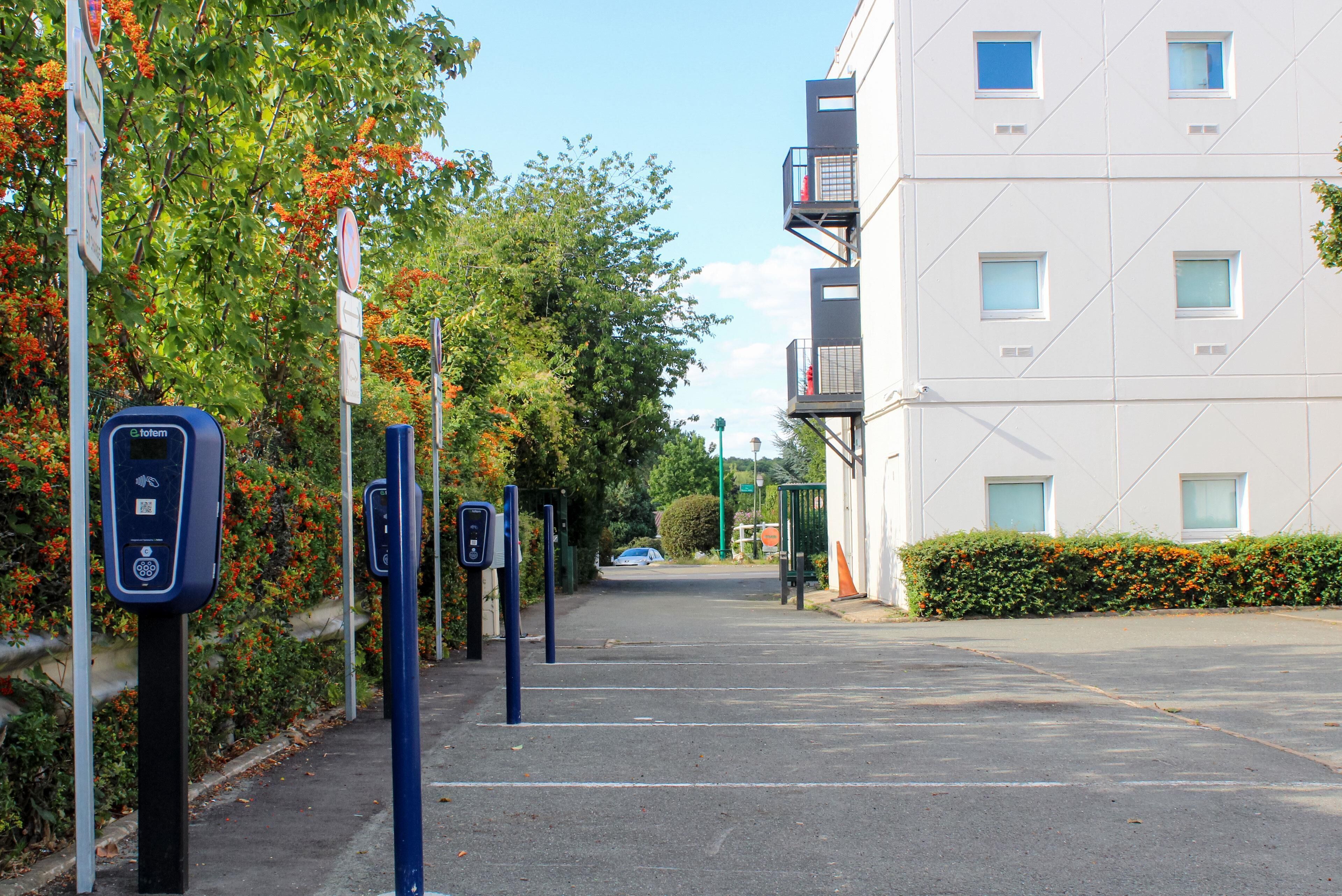 Un área de estacionamiento con estaciones de carga para vehículos eléctricos, cielo despejado y un edificio con balcones.