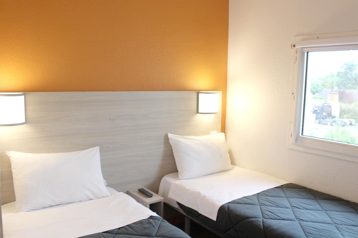 A hotel room with two beds, each with white bedding and a dark-colored quilt. There are two bedside tables with lamps, and a window on the right side.