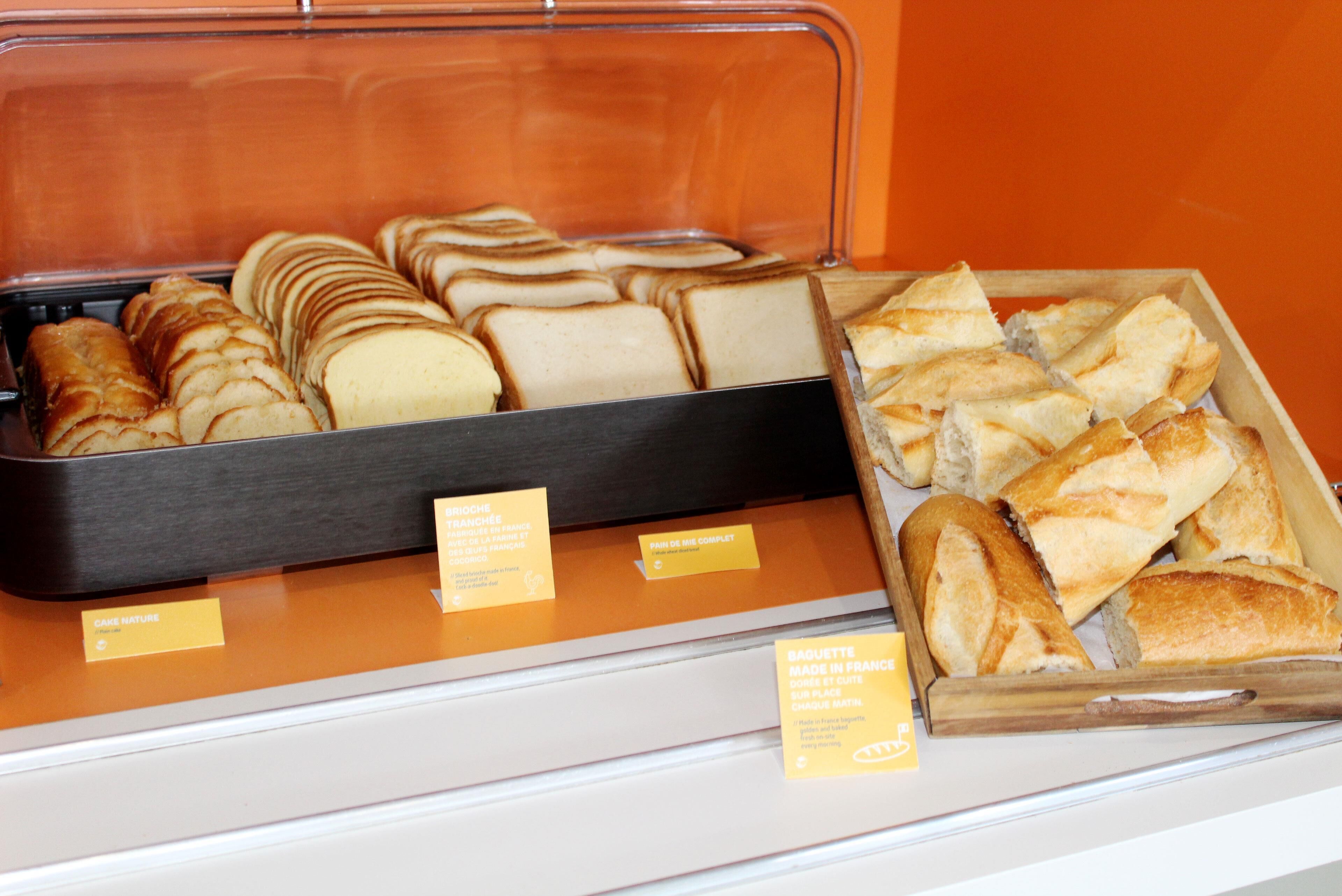 A display case with various types of bread, including baguettes and sliced bread, with labels indicating their origin and type.