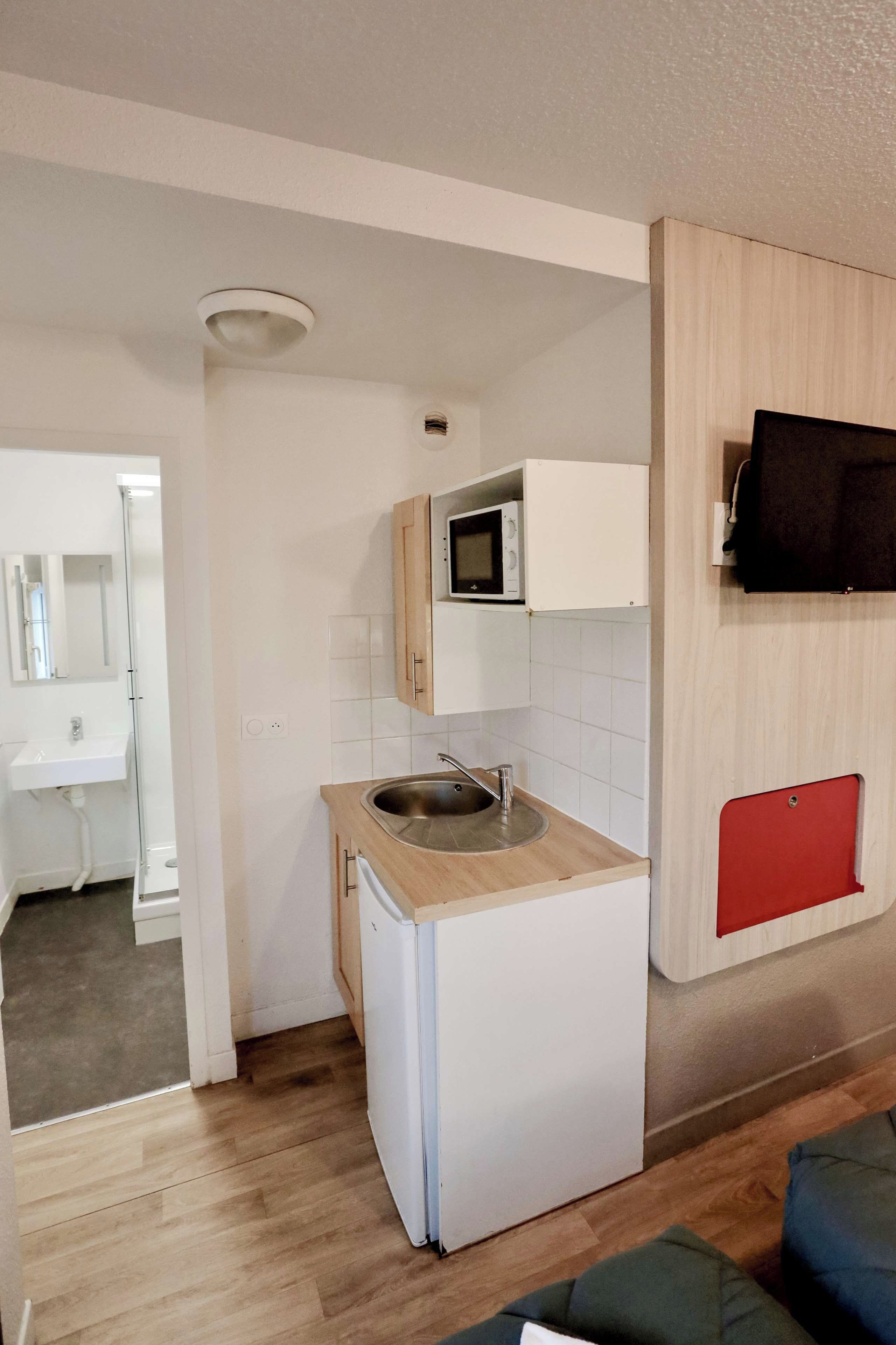 A compact kitchen with a microwave, sink, and a small refrigerator. A red cabinet is visible on the right, and a wooden cabinet is above the microwave.