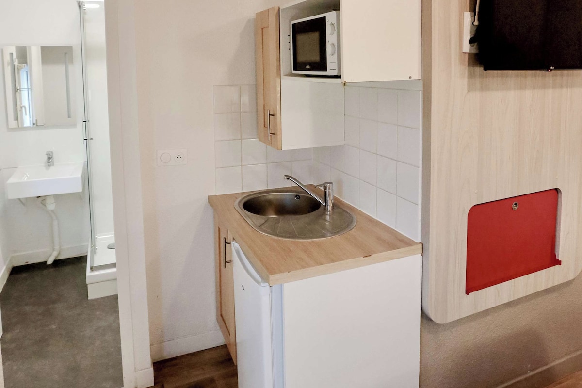 A compact kitchen with a microwave, sink, and a small refrigerator. A red cabinet is visible on the right, and a wooden cabinet is above the microwave.