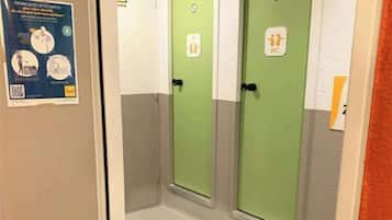 Un couloir avec deux portes vertes, l’une portant l’inscription « WC » et l’autre l’inscription « T ». Une affiche est accrochée au mur, sur la gauche.