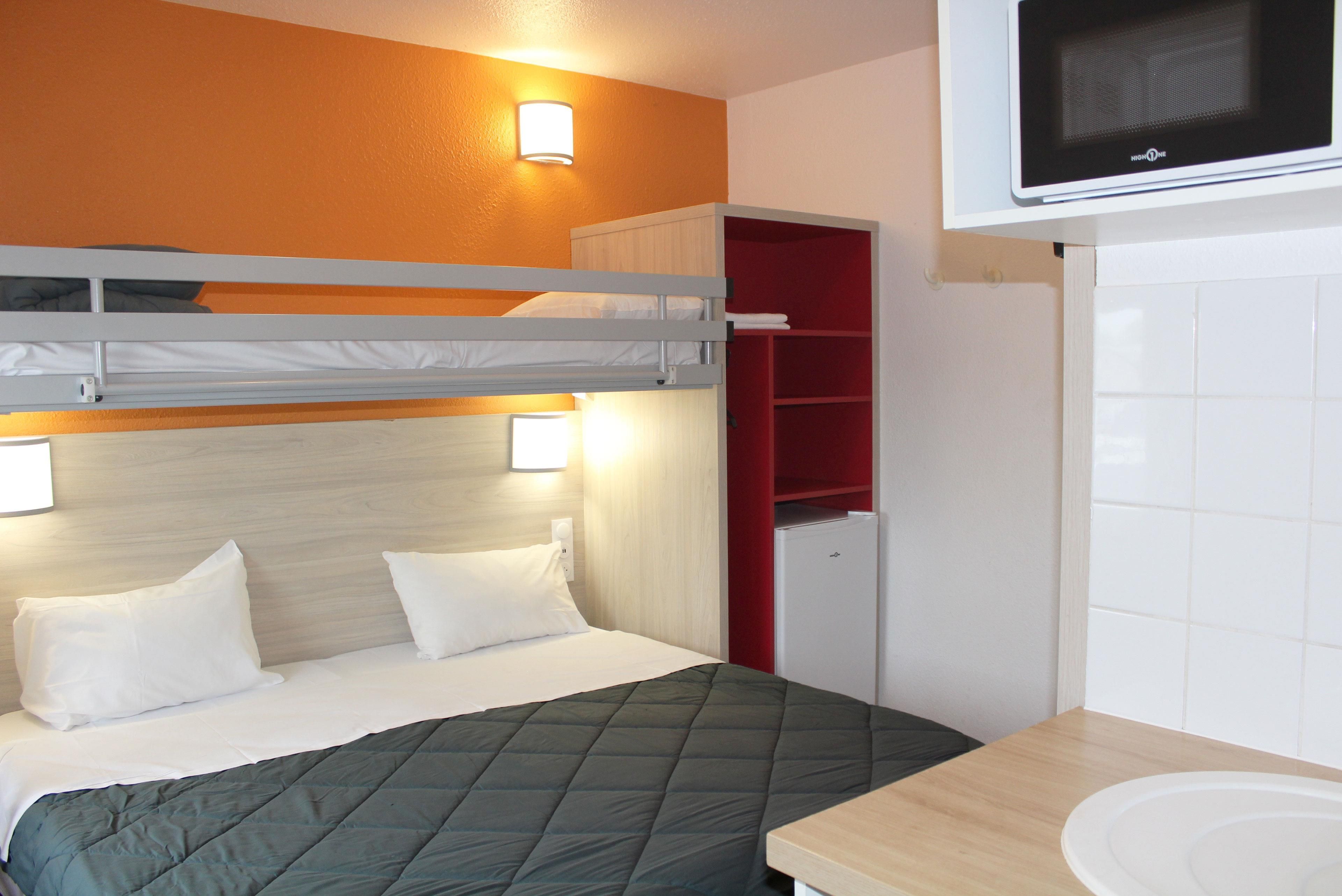 Een stapelbed met een groene quilt, een witte nachtkastje, een magnetron boven het bed en een rode opbergkast.