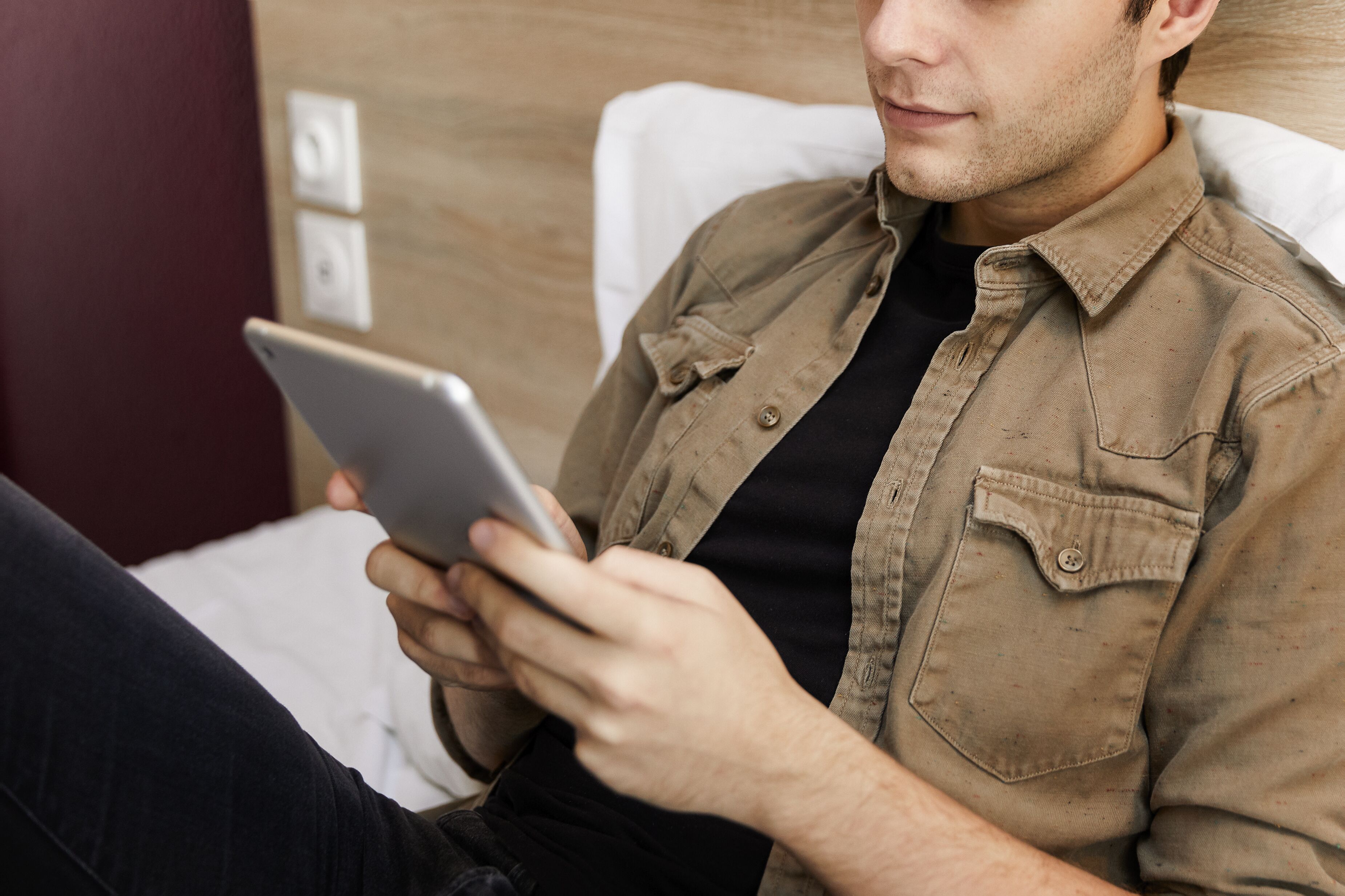 A person sitting on a bed with a tablet, wearing a brown shirt and black pants.