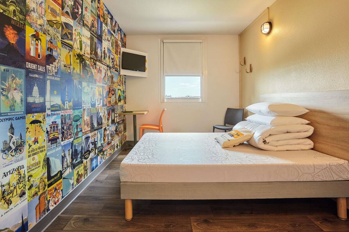 A hotel room with a bed, a desk, a chair, a TV, and a wall covered with travel posters.