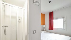 A hotel room with a shower, a bed, a window with a red curtain, and a wall-mounted light fixture. - Premiere Classe Angers Est - St Barthelemy D'Anjou (Saint-Barthelemy-d'Anjou)