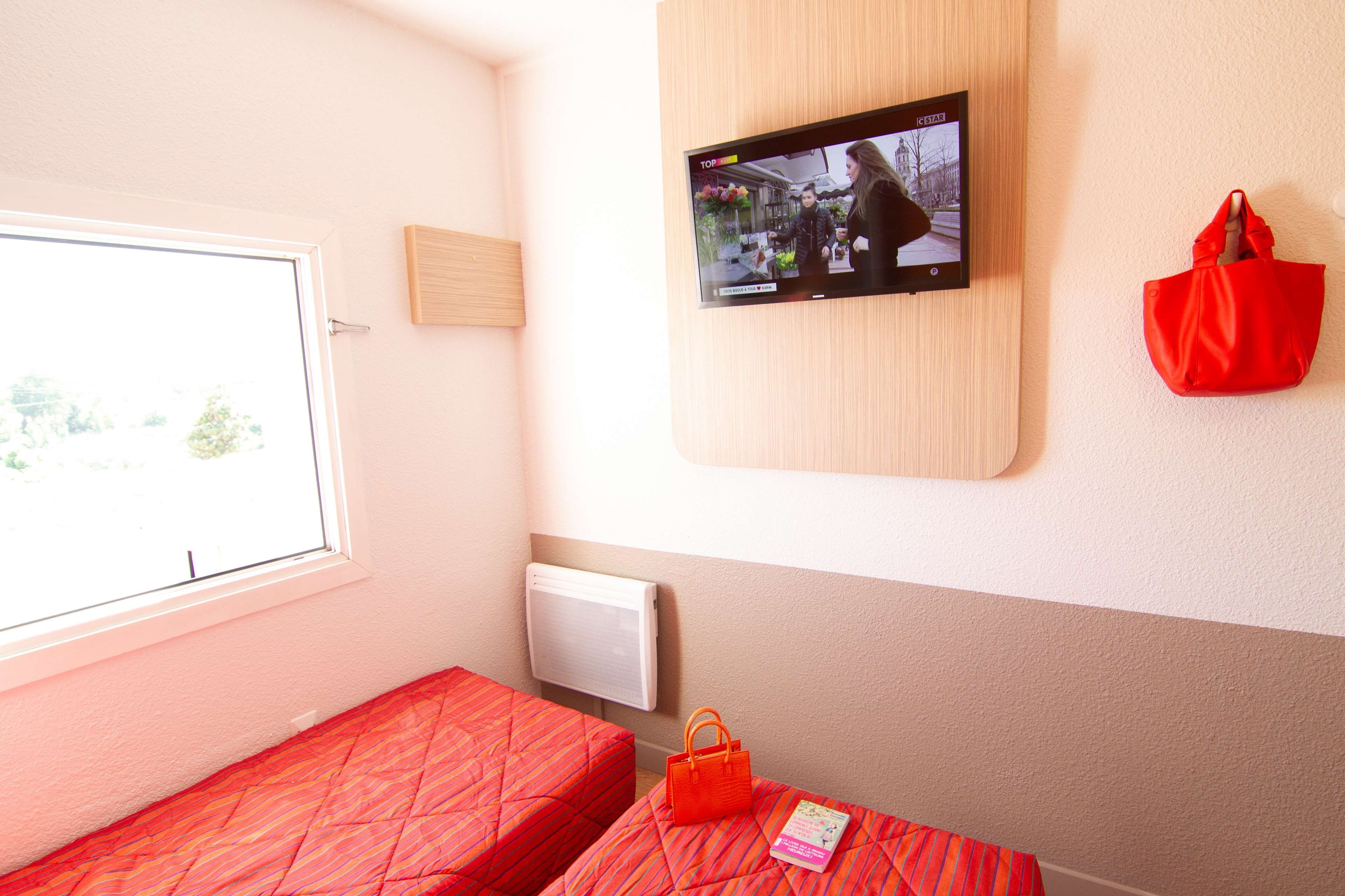 Habitación de hotel con dos camas, un televisor montado en la pared, una ventana y un bolso rojo colgado en la pared.