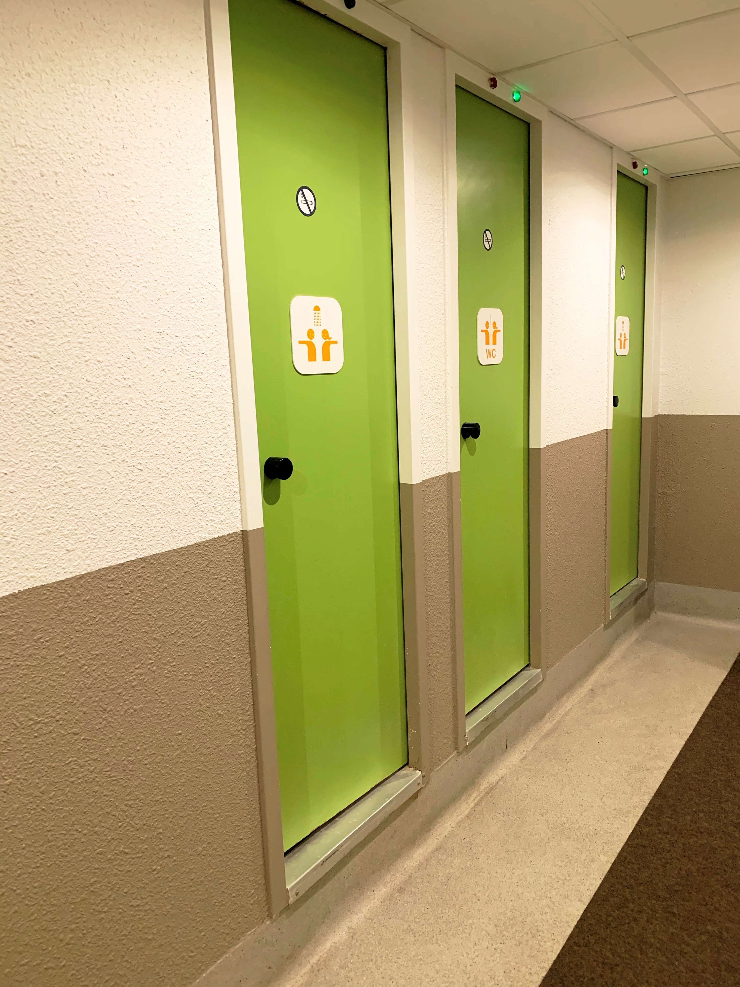 Ein Korridor mit grünen Türen, jede mit einem weißen Schild, das auf eine Herrentoilette hinweist.