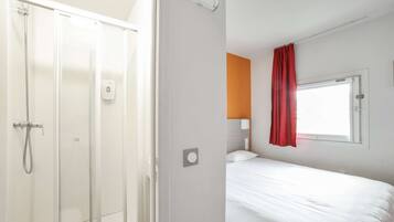 Une chambre d’hôtel avec une douche, un lit, une fenêtre avec un rideau rouge et un luminaire fixé au mur.