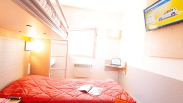 Una habitación de hotel con una cama, una ventana, un ordenador portátil sobre una estantería y un televisor mostrando un anuncio de coche.