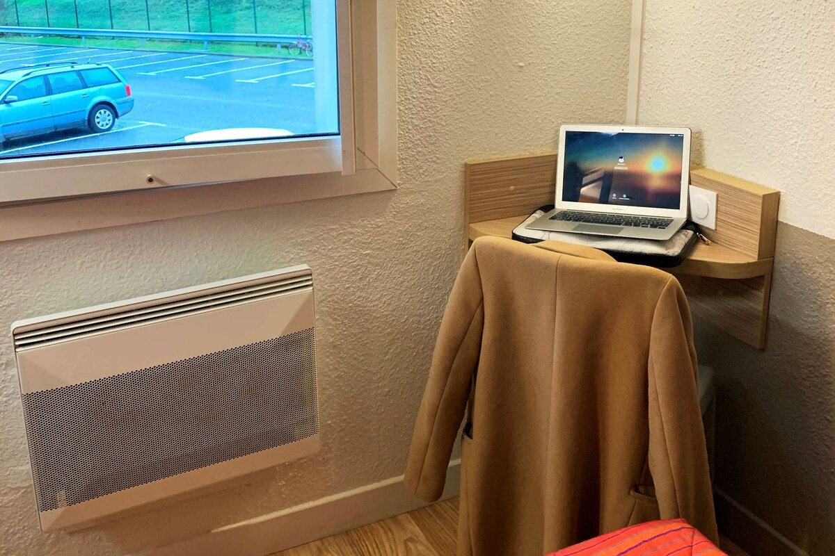 Une chambre d’hôtel avec une fenêtre, un ordinateur portable posé sur une petite table, un radiateur et un lit recouvert d’une couverture à rayures rouges.