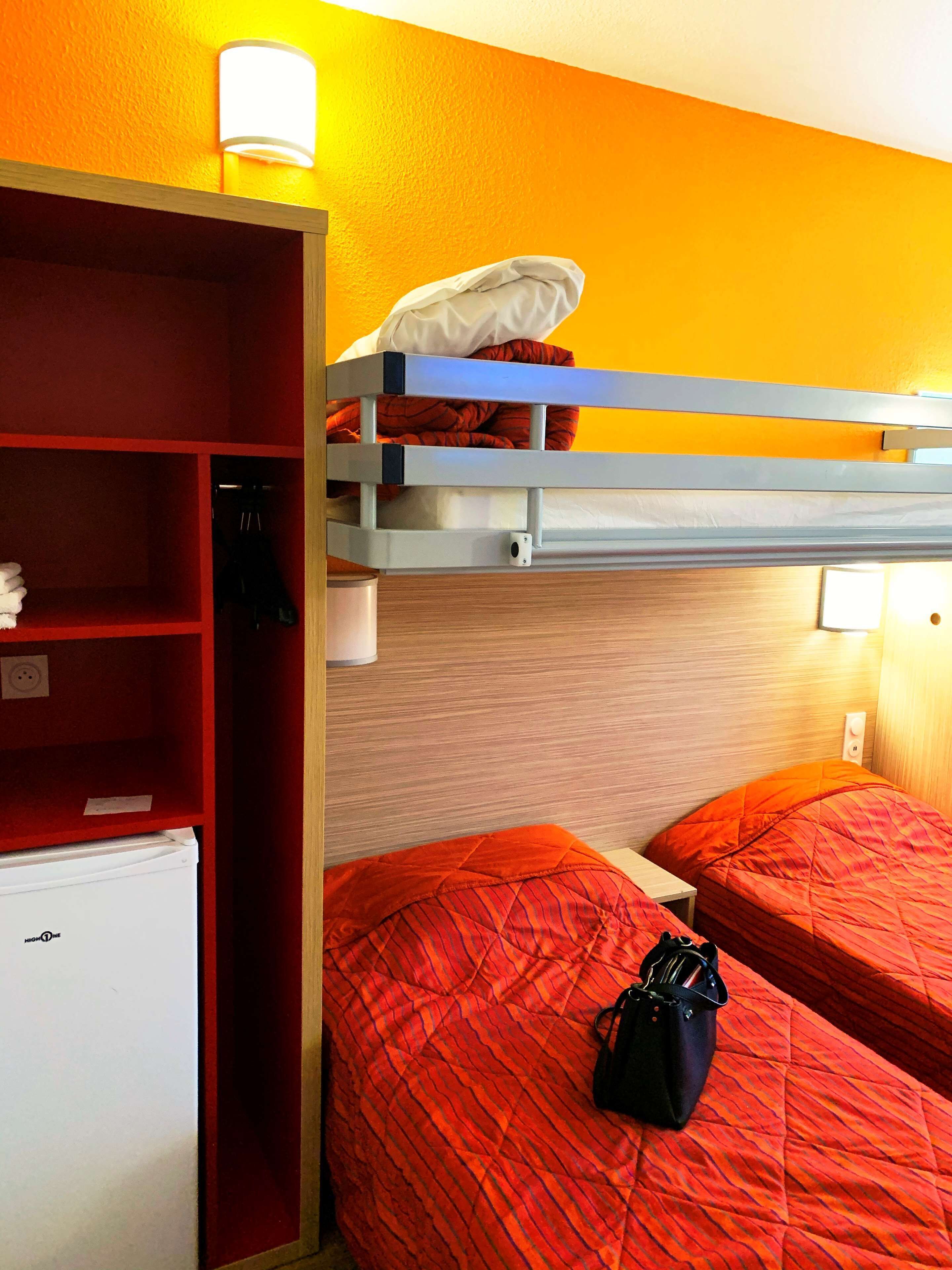 A hotel room with a bunk bed, a small refrigerator, a shelf with a lamp, and a suitcase on the bed.