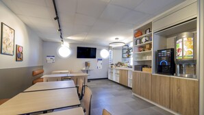 A modern cafe with wooden tables and chairs, a coffee machine, and a fruit juice dispenser. - ibis budget Forbach Centre (Forbach)