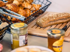 A variety of baked goods including croissants and a loaf of bread with seeds, alongside jars of honey and a knife on a wooden cutting board.