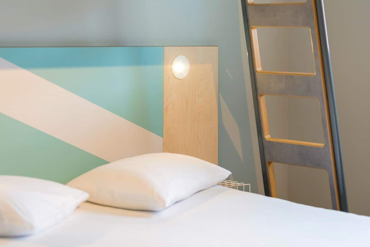 Una cama con ropa de cama y almohadas blancas, un cabecero de madera y una escalera metálica apoyada en la pared.