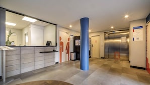 A reception area with a counter, a blue pillar, an elevator, and a restroom symbol on the wall. - ibis budget Forbach Centre (Forbach)