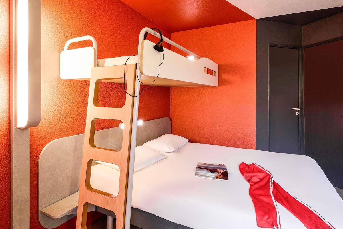 A bunk bed with a ladder, a bedside table, a bed with white bedding, a door, and a wall-mounted light fixture.