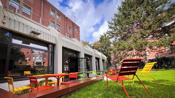 Un espace extérieur aménagé avec des chaises et des tables aux couleurs vives, une pelouse verdoyante et un bâtiment doté de grandes fenêtres et d’une façade en briques.
