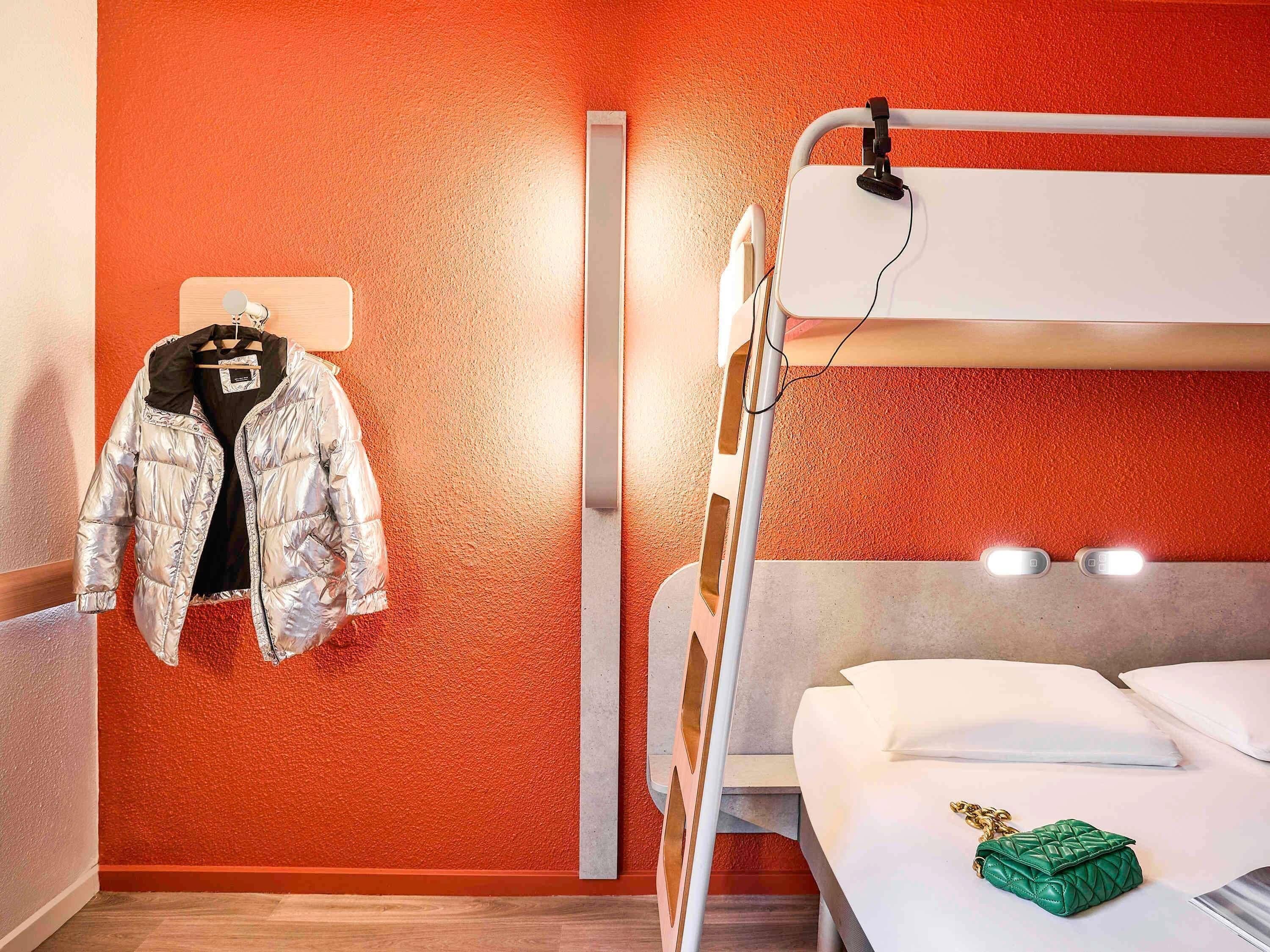 Un lit superposé avec un matelas et des oreillers blancs, un porte-manteau suspendu avec une veste métallique, et un sac à main vert posé sur le lit.