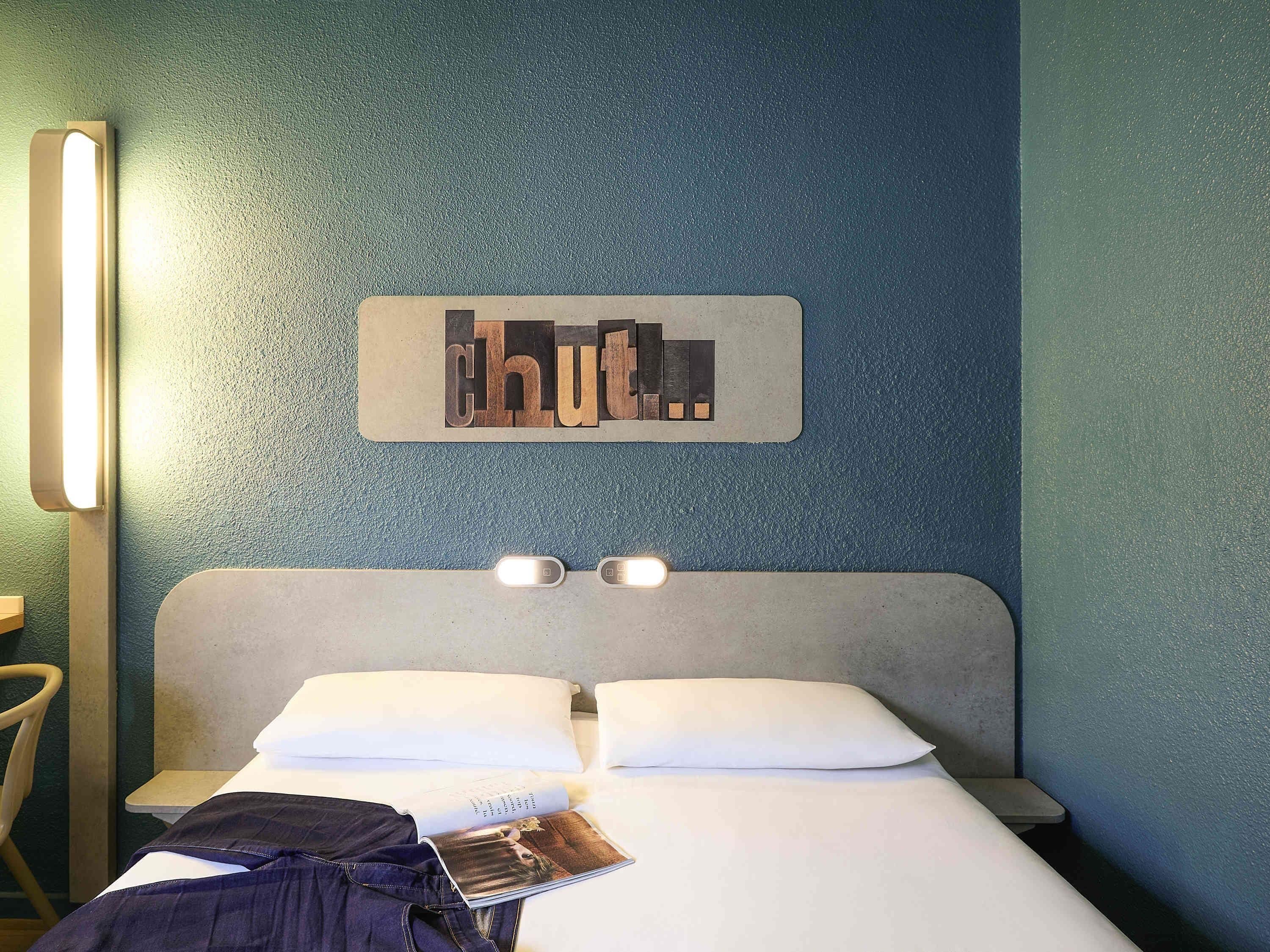 A hotel room with a bed, pillows, a nightstand, a lamp, and a sign on the wall.