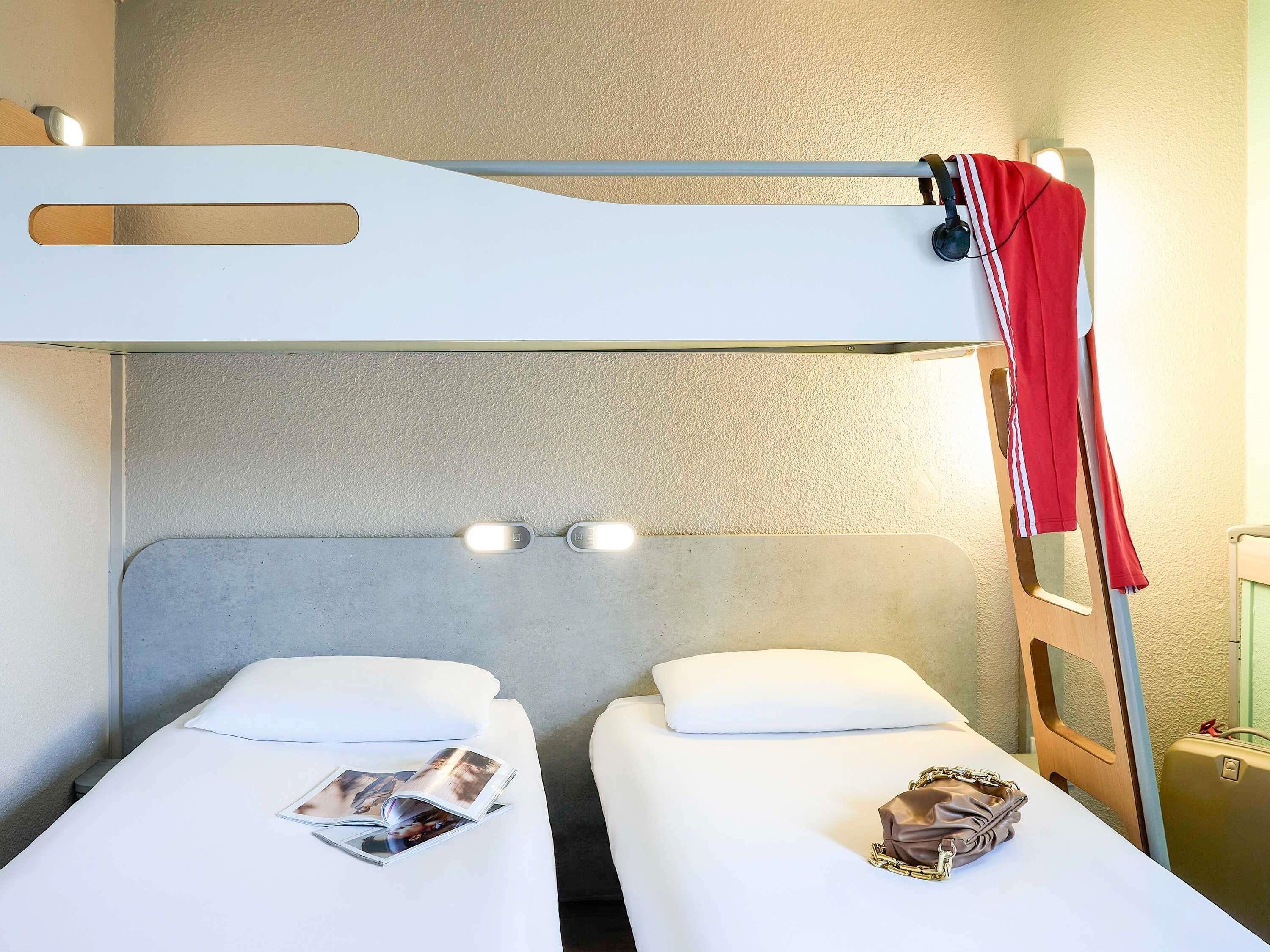 Une chambre avec un lit superposé, deux lits, une échelle, une valise et un magazine posé sur l’un des lits.