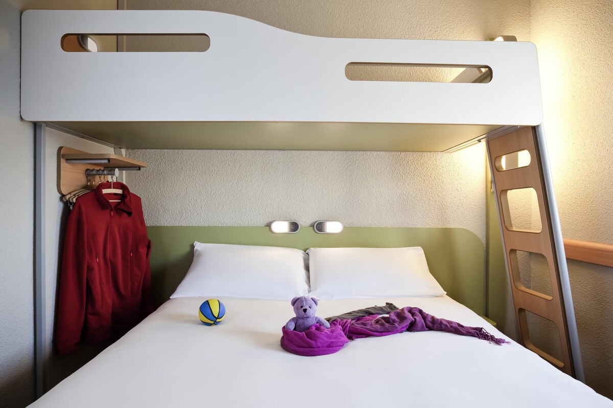 Een stapelbed met hoofdbord, een nachtkastje met lamp, een rood jasje hangend, een paars dekbed en een strandbal.