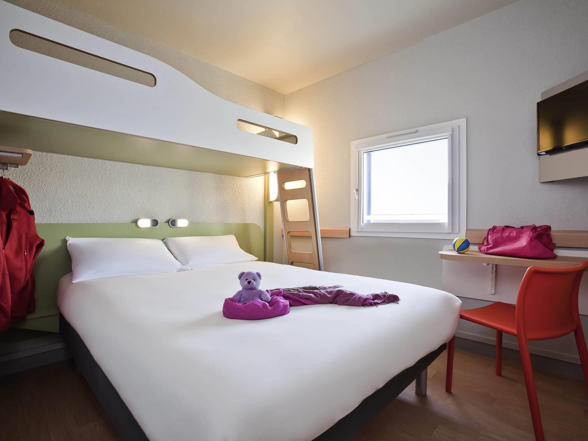 A hotel room with a bed, a desk, a chair, a window, and a teddy bear on the bed.