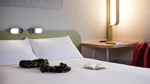 Ein ordentlich gemachtes Bett mit weißen Kissen und einem schwarzen, gemusterten Gegenstand darauf. Auf dem Bett liegt ein aufgeschlagenes Buch. Eine Lampe steht auf einem Nachttisch neben dem Bett.
