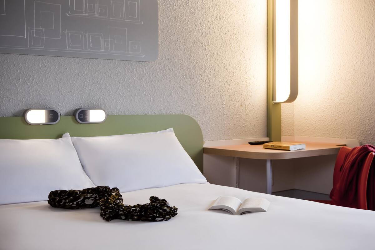 Un lit bien fait, avec des oreillers blancs et un objet noir à motifs posé dessus. Un livre est ouvert sur le lit. Une lampe se trouve sur une table de chevet, à côté du lit.