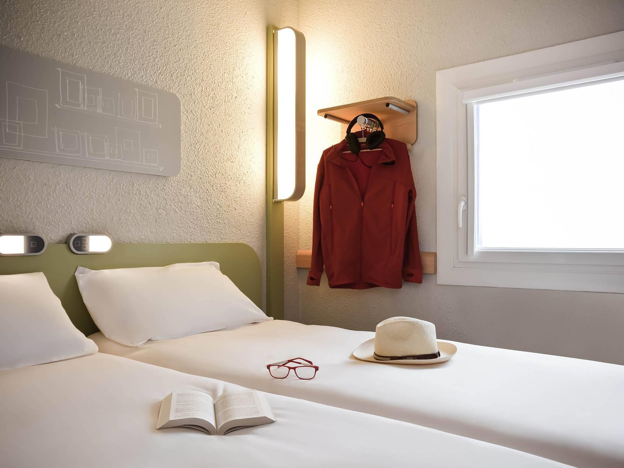 A hotel room with a bed, a window, a wall-mounted light, and a coat rack with a red coat.