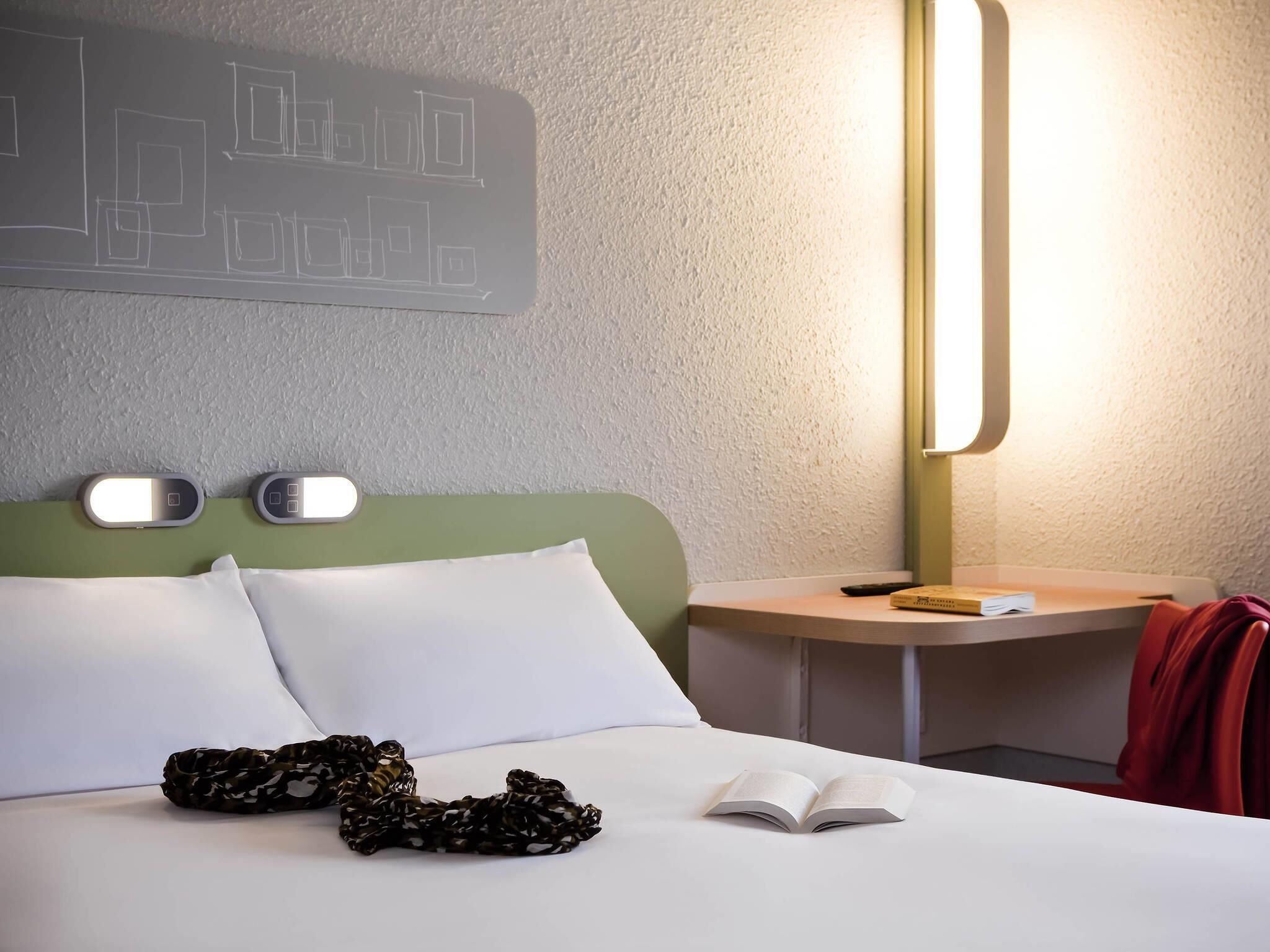 A hotel room with a bed, pillows, a blanket, an open book, a bedside table with a lamp, and a wall-mounted light fixture.