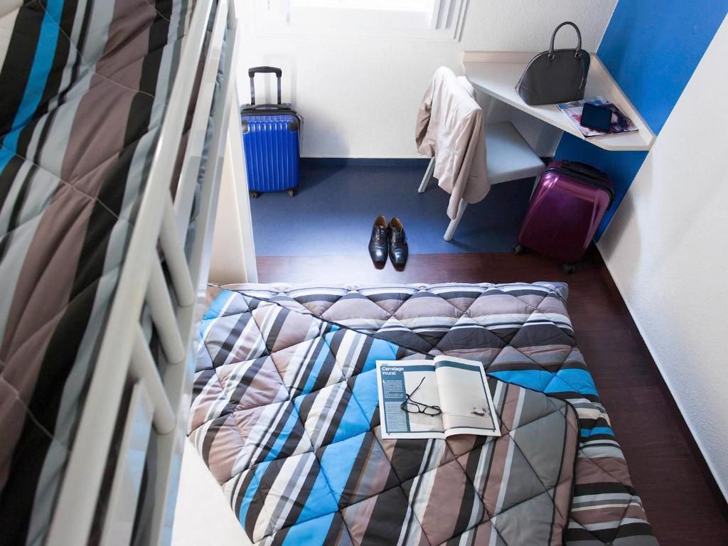 Una habitación de hotel con una cama, una maleta, un bolso y una revista sobre la cama.