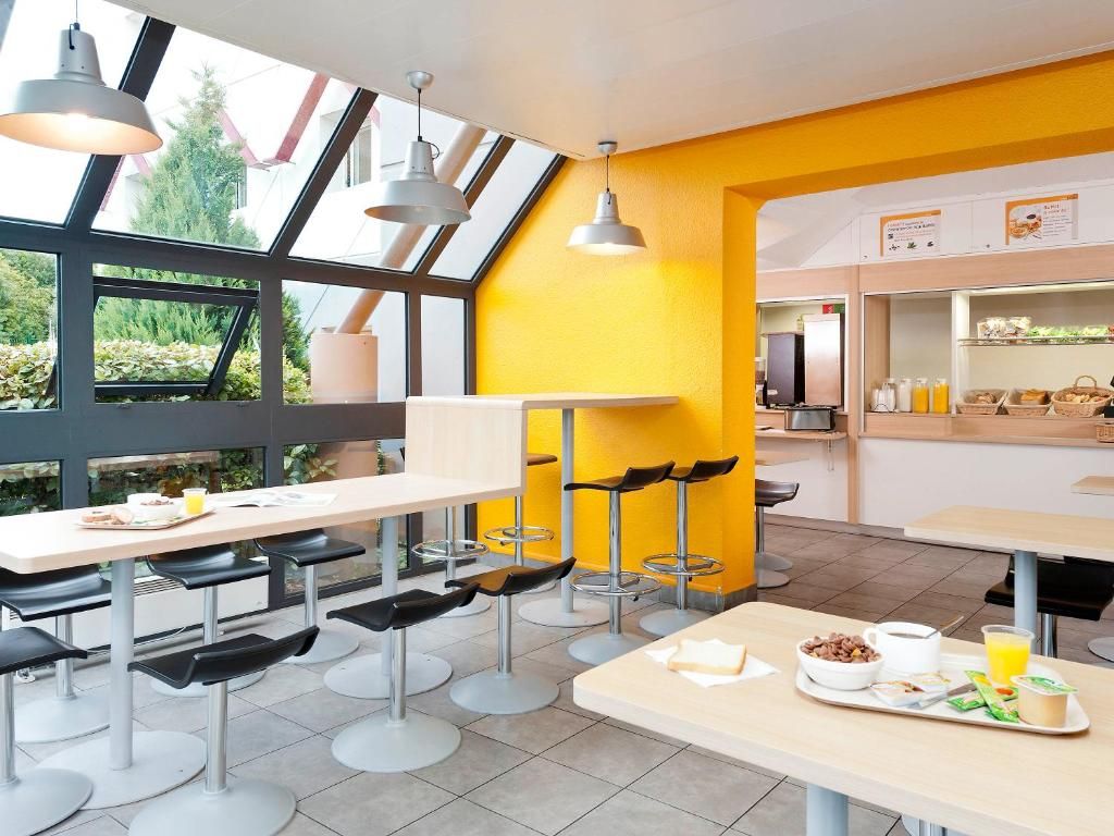 Un café moderne avec de hautes tables et des tabourets de bar, un mur d’accent jaune vif et une vue dégagée grâce à de grandes fenêtres.