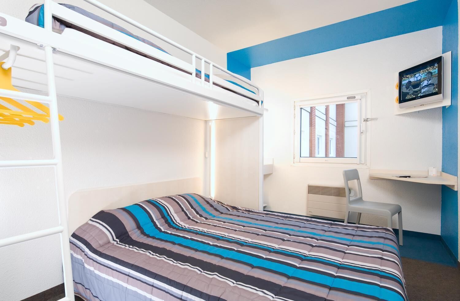 Een slaapkamer met een stapelbed, een beddegoed met strepen, een bureau met stoel, een televisie en een raam met uitzicht op gebouwen.
