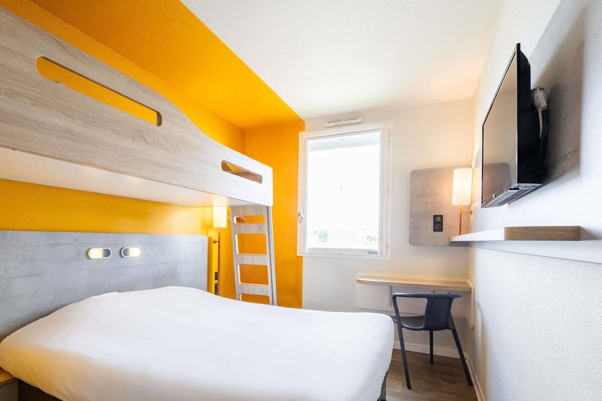 A hotel room with a bunk bed, a desk, a chair, a TV, and a window.