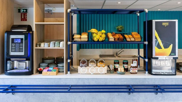 Una estación de desayuno autoservicio con una máquina de café, una vitrina de pan y diferentes condimentos.