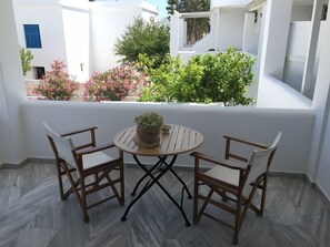 A small outdoor seating area with a round table and two chairs, a potted plant on the table, and a view of a white building with balconies and greenery. - CAELI PAROS by Kostas and Ioanna Studios (Paros)
