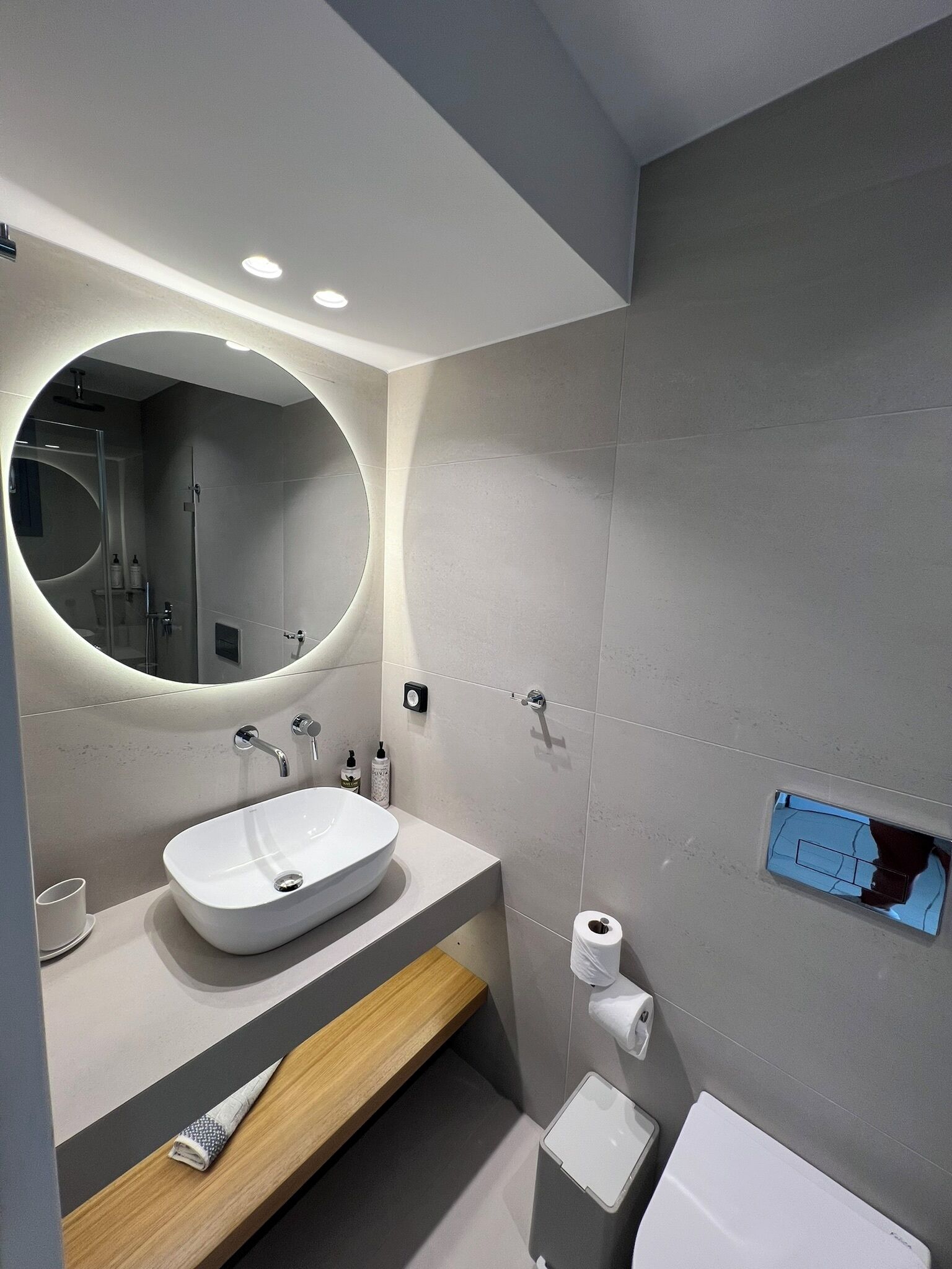 A modern bathroom with a large oval mirror, a white sink, and a toilet.