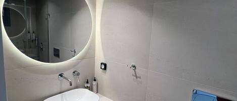 A modern bathroom with a large oval mirror, a white sink, and a toilet.