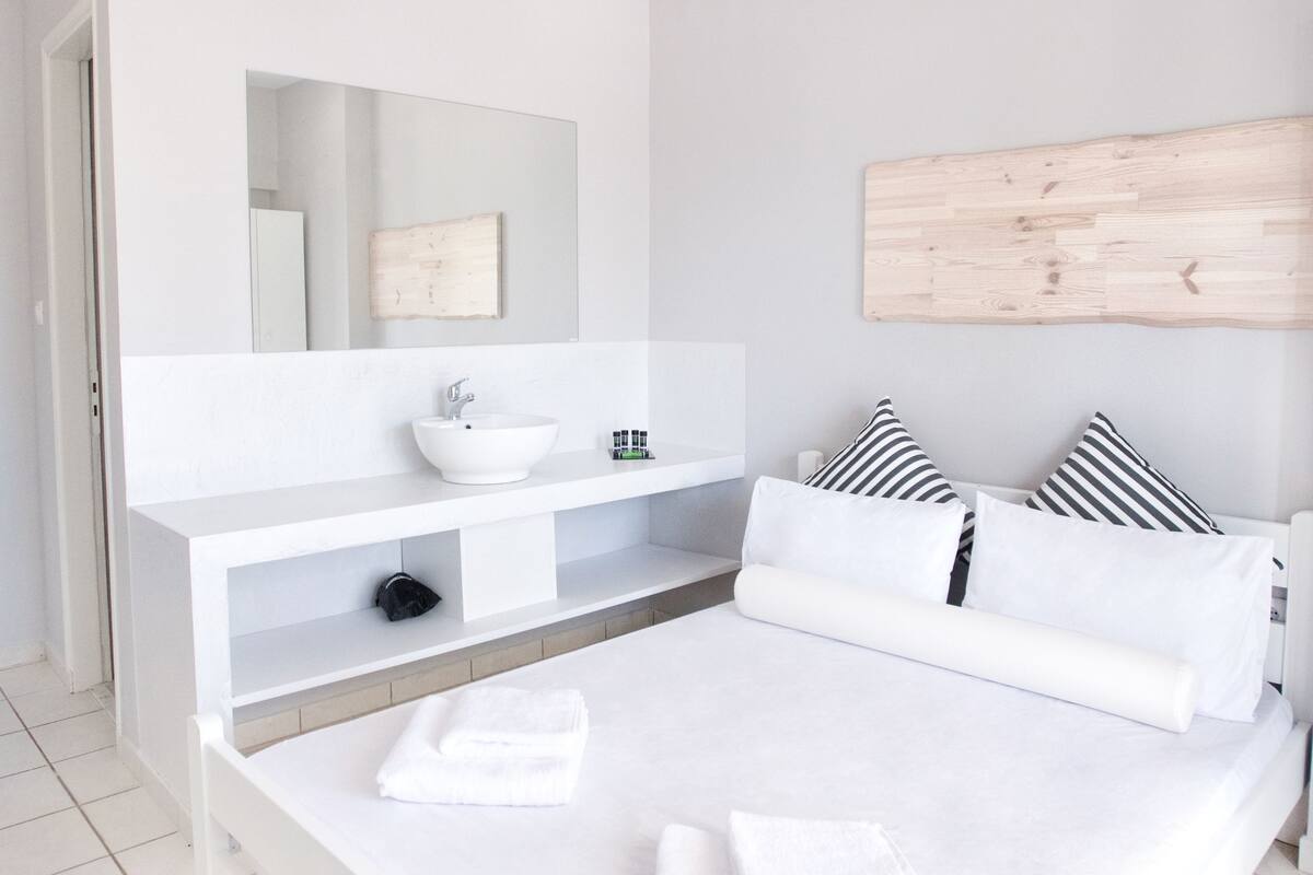 Un dormitorio moderno con cama blanca, un espejo grande, un tocador blanco e iluminación colgante.