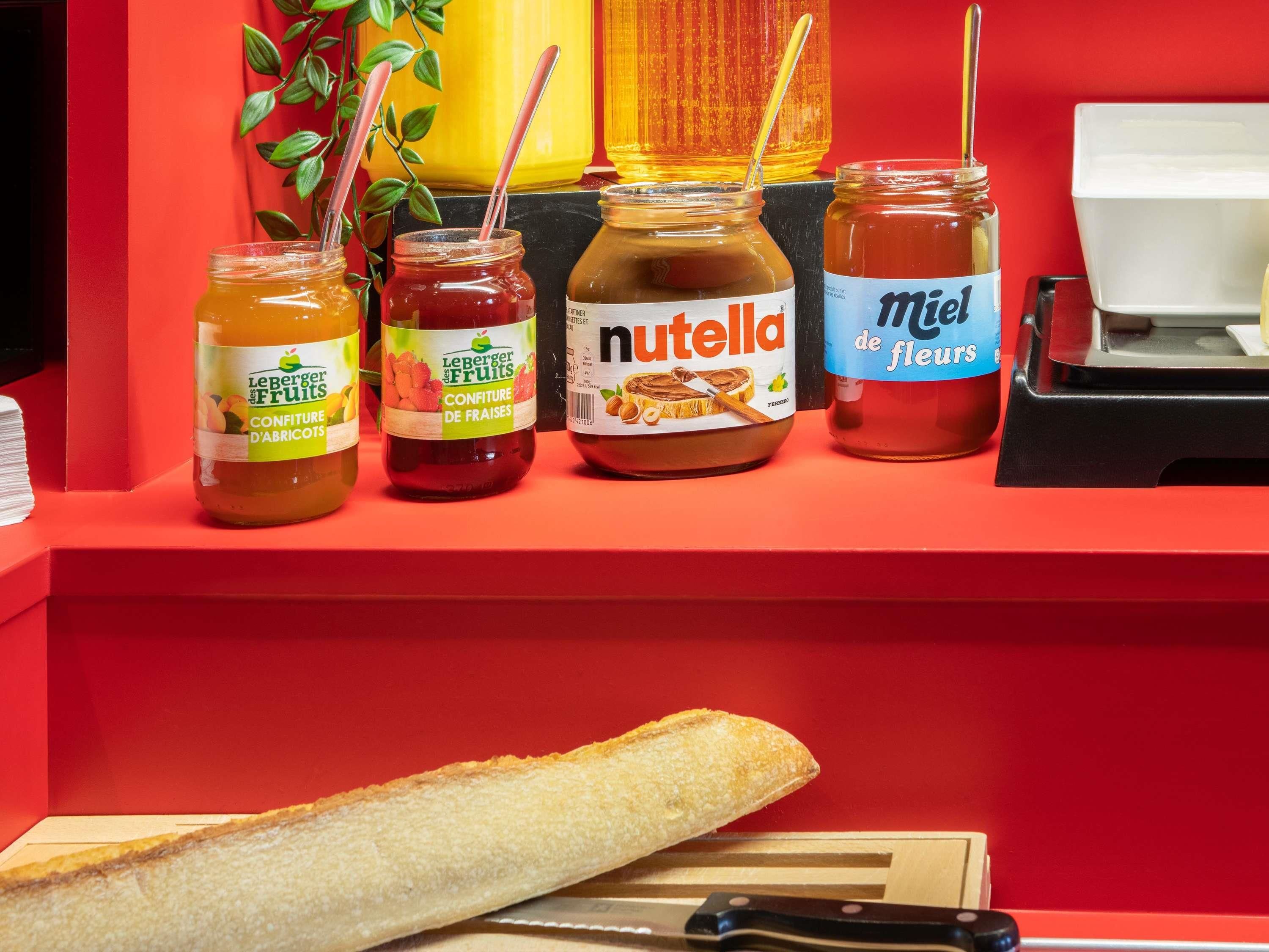 Une variété d’aliments sont exposés, notamment des pots de confitures de fruits et une baguette, le tout présenté sur un comptoir rouge.