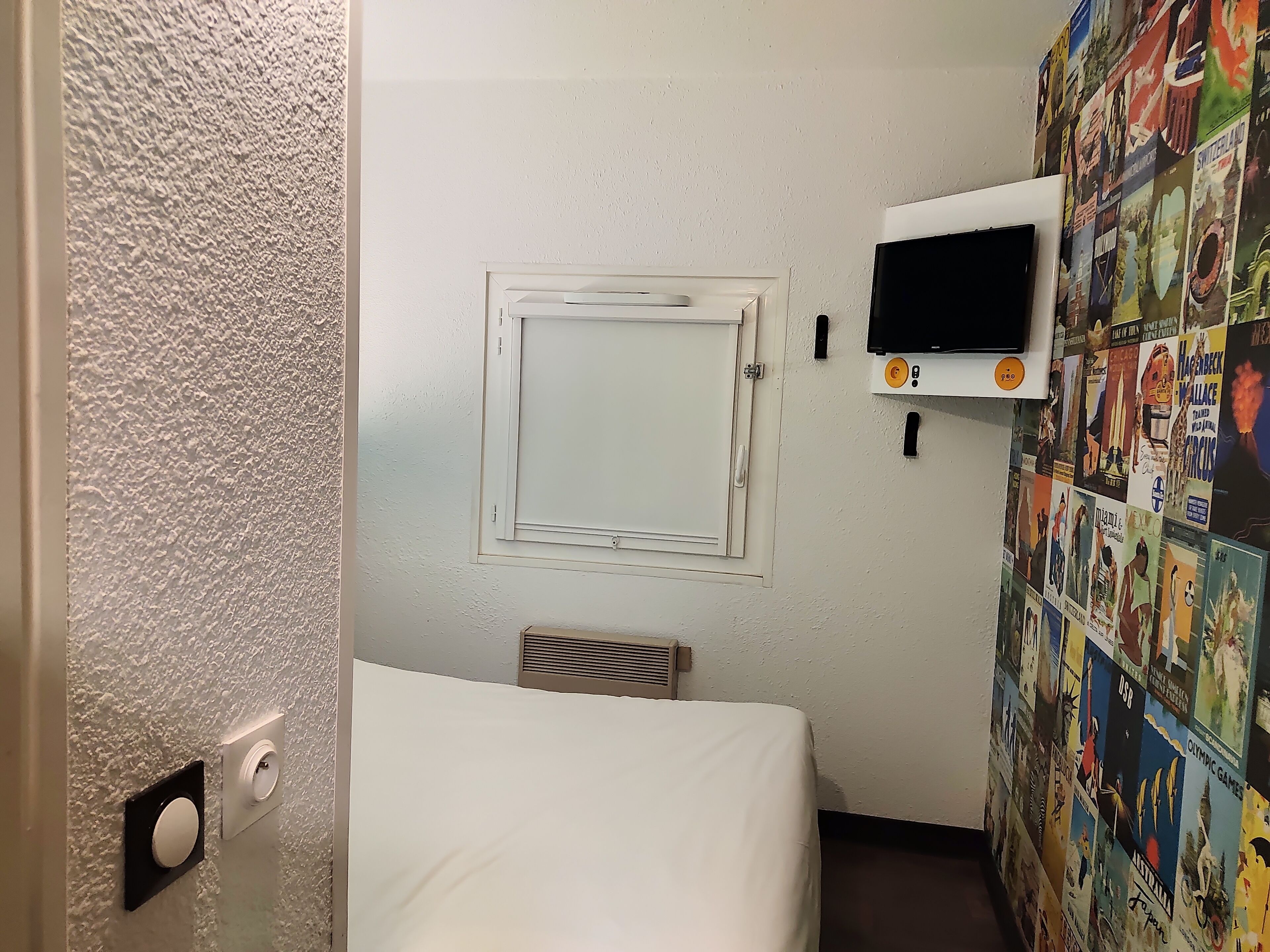 Ett litet hotellrum med en säng, en tv, ett fönster med utsikt och en vägg som är dekorerad med olika affischer.