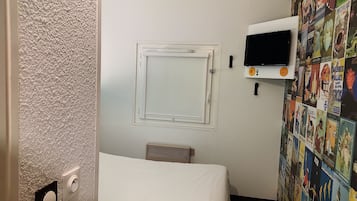 A small hotel room with a bed, a TV, a window with a view of the outside, and a wall decorated with various posters.
