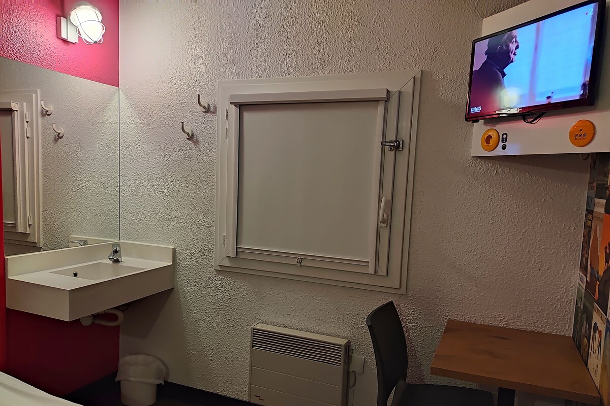 A hotel room with a white sink, a mirror, a television mounted on the wall, a window with a white frame, and a small table with a chair.